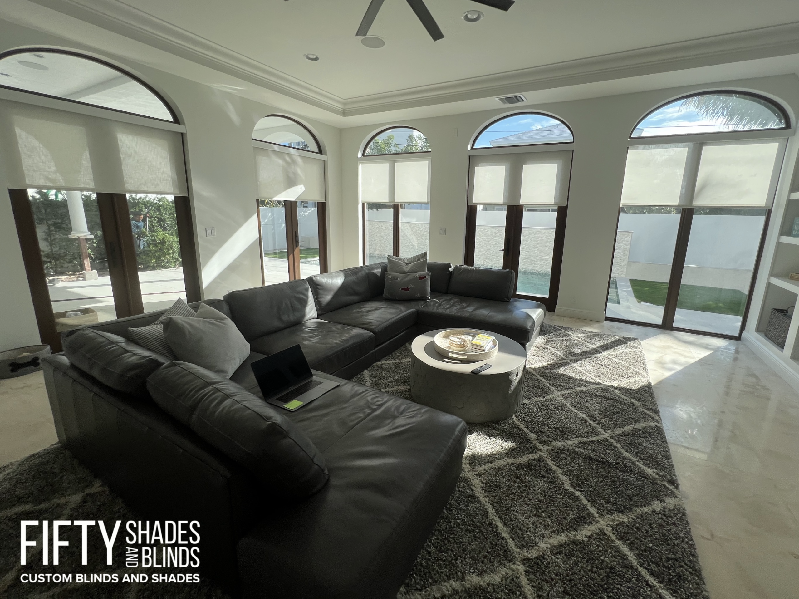 Bright living room with tall arched windows covered by white roller blinds, a gray sectional sofa, and a round coffee table; branding text in the corner reads 'FIFTY SHADES'.
