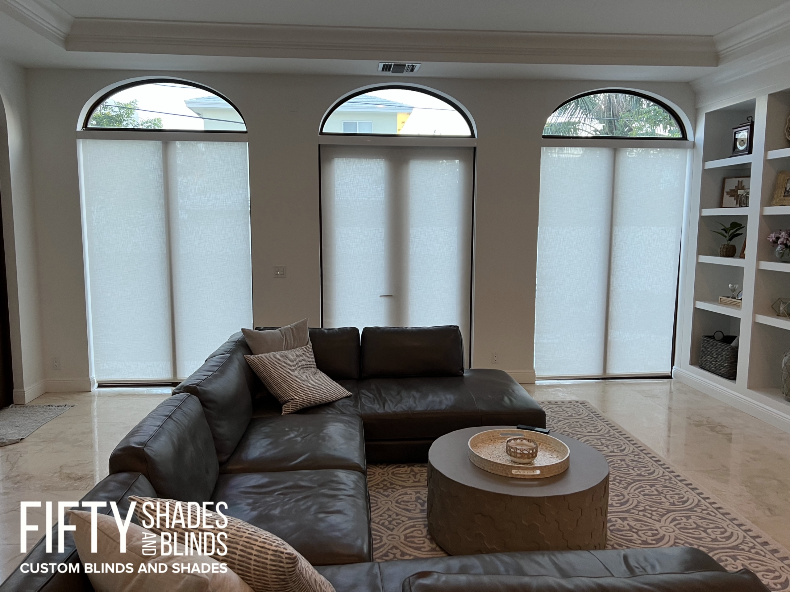 Living room showing tall arched windows with light roller shades, a gray sectional sofa, and a round woven coffee table; built-in shelves line the right wall. Watermark reads 'FIFTY SHADES AND BLINDS'.