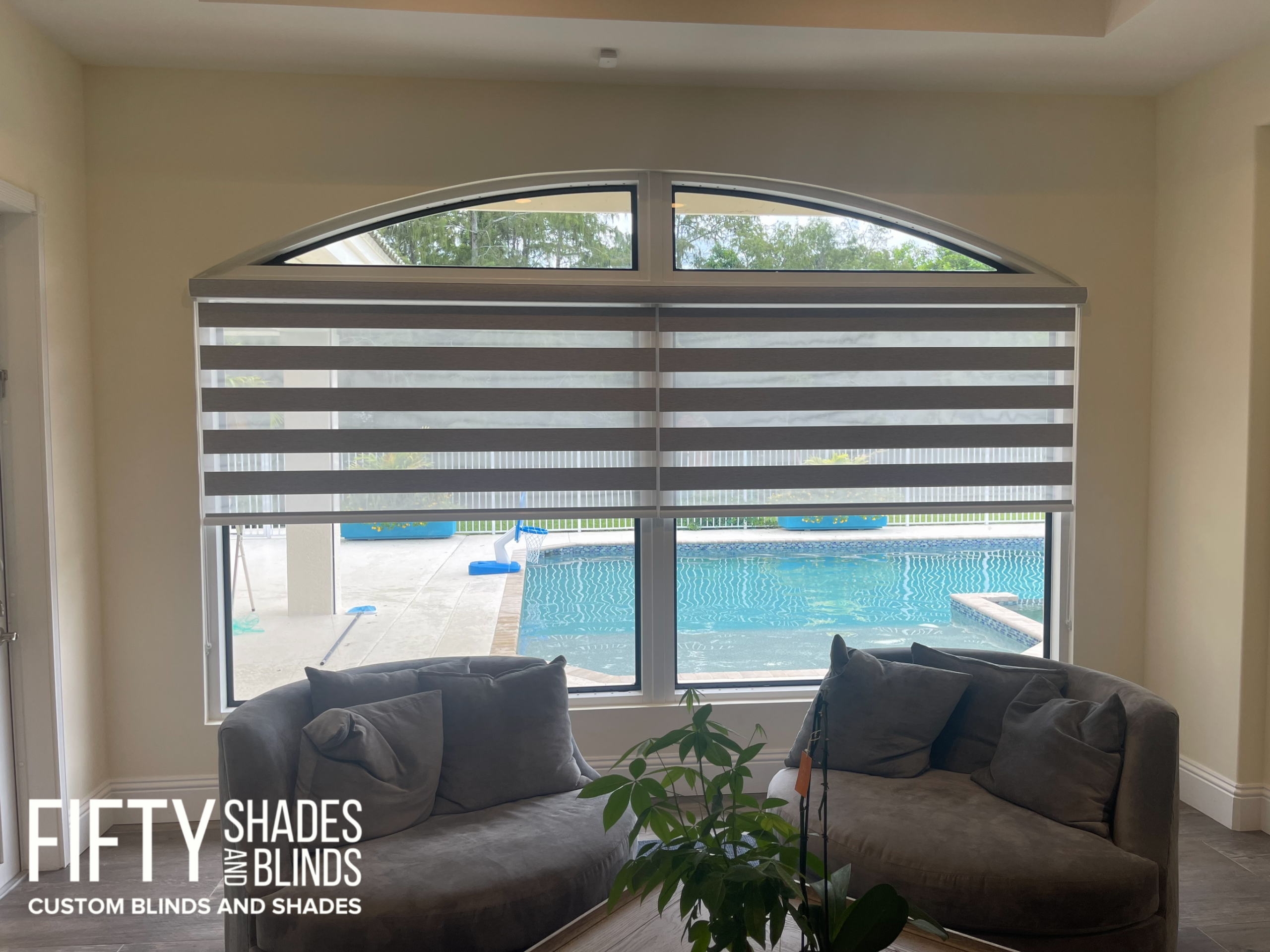Living room with a large arched window and striped dual-layer blinds; outdoor pool area visible through the window, with branding text overlay reading 'LIFTY SHADES AND BLINDS'
