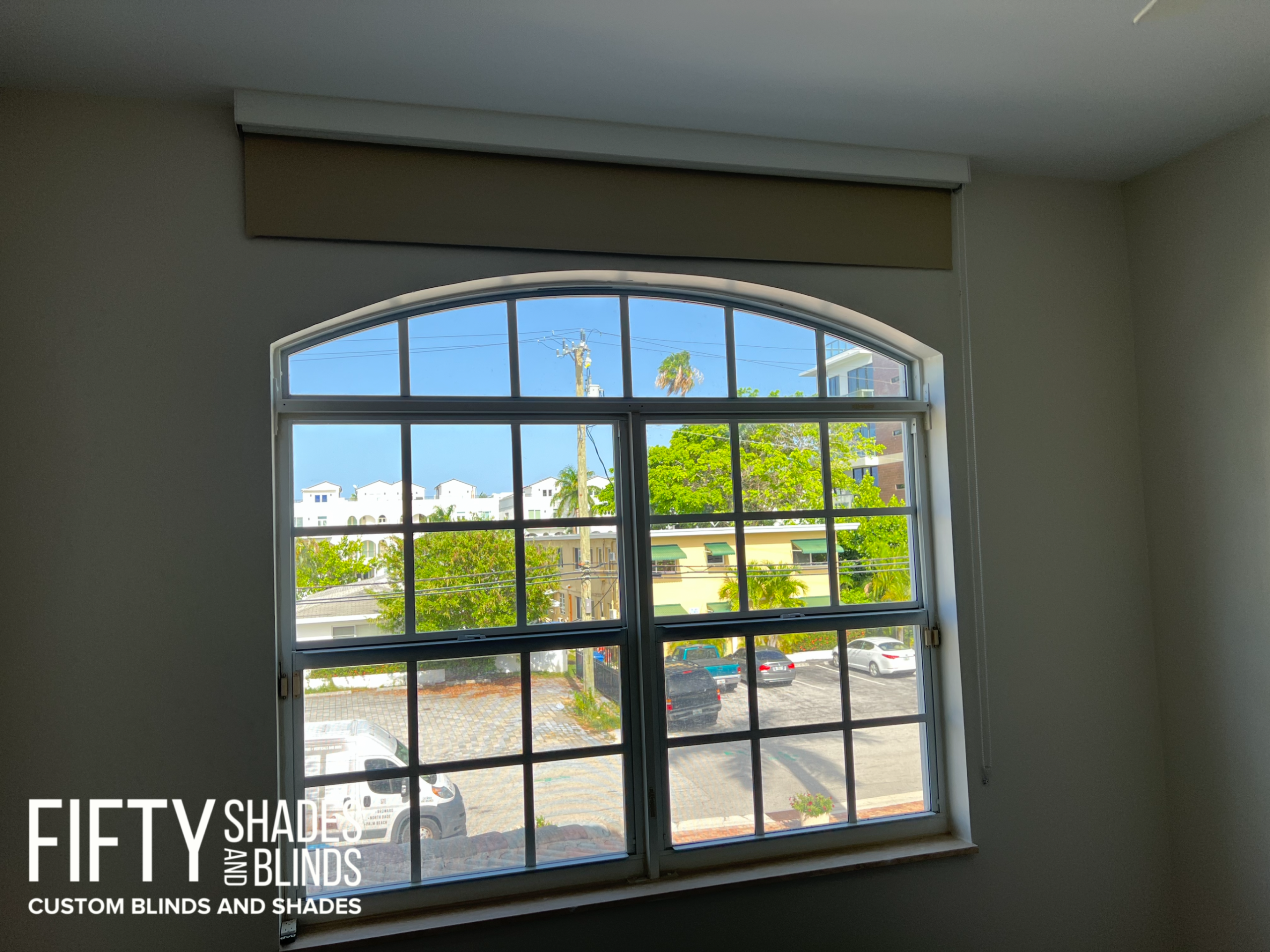 Interior of a room showing a large arched window with a beige roller shade rolled up at the top; view of street and buildings outside.