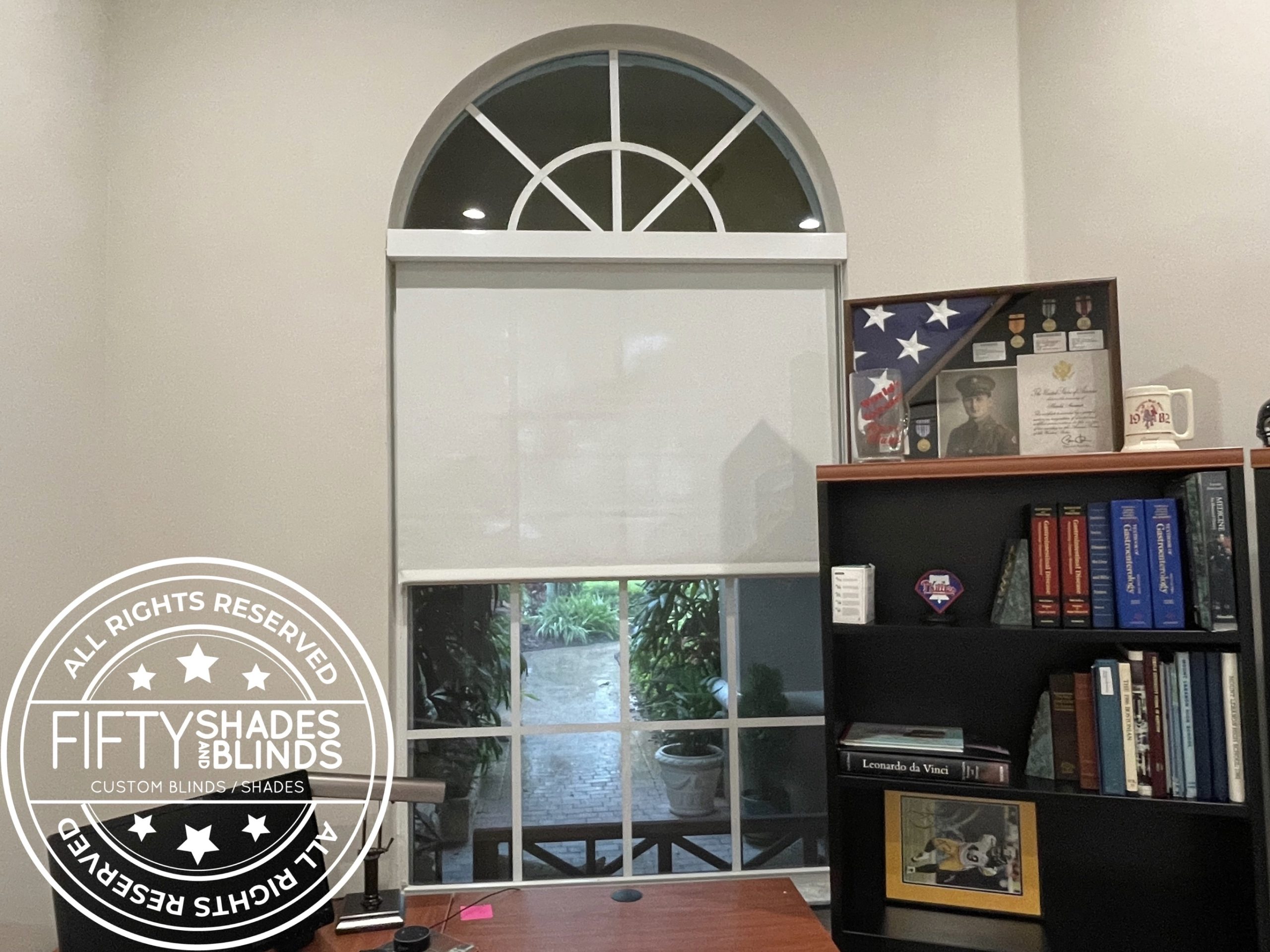 Office blinds showroom with an arched window and white blinds, a large circular watermark logo on the left, and shelves of books and awards nearby.