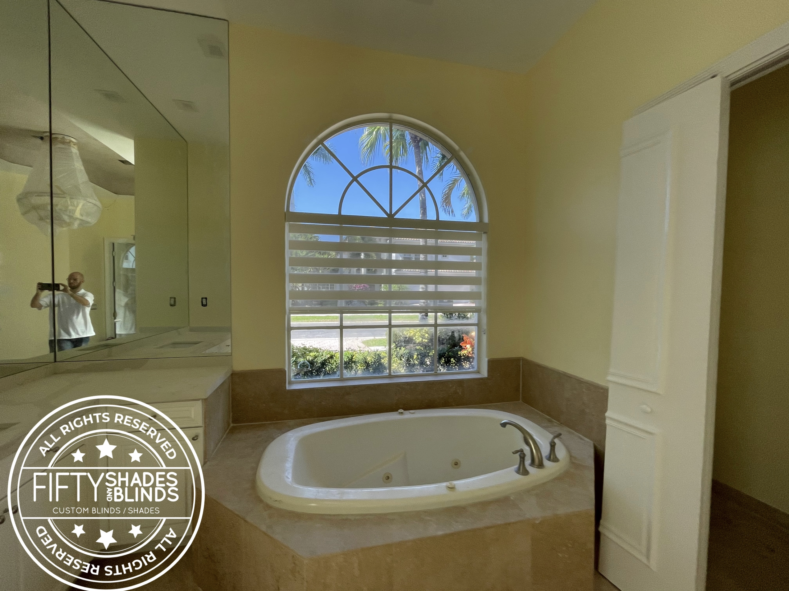 Bathroom with built-in oval tub, beige tile surround, arched window with horizontal blinds, reflecting a person taking a photo in a large mirror.